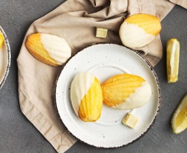 Lemon Madeleines dipped in white chocolate on a white plate on a cloth napkin.