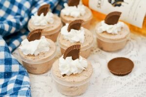 Skrewball Whiskey Pudding shots with whipped cream topping and Peanut butter cup garnish on a table with blue checked cloth.