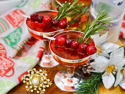 Poinsettia cocktail with vodka with whole cranberry skewer and fresh rosemary sprig for garnish, sitting on a wood table.