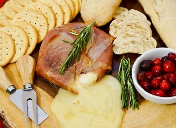 Prosciutto Wrapped Brie baked and served on wood board with crackers and bread around it.