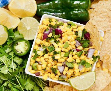 Chipotle Corn Salsa in a bowl surrounded with cut limes, cilantro, a jalapeno pepper and tortilla chips for dunking.