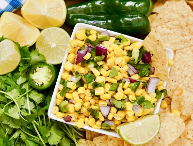 Chipotle Corn Salsa in a bowl surrounded with cut limes, cilantro, a jalapeno pepper and tortilla chips for dunking.