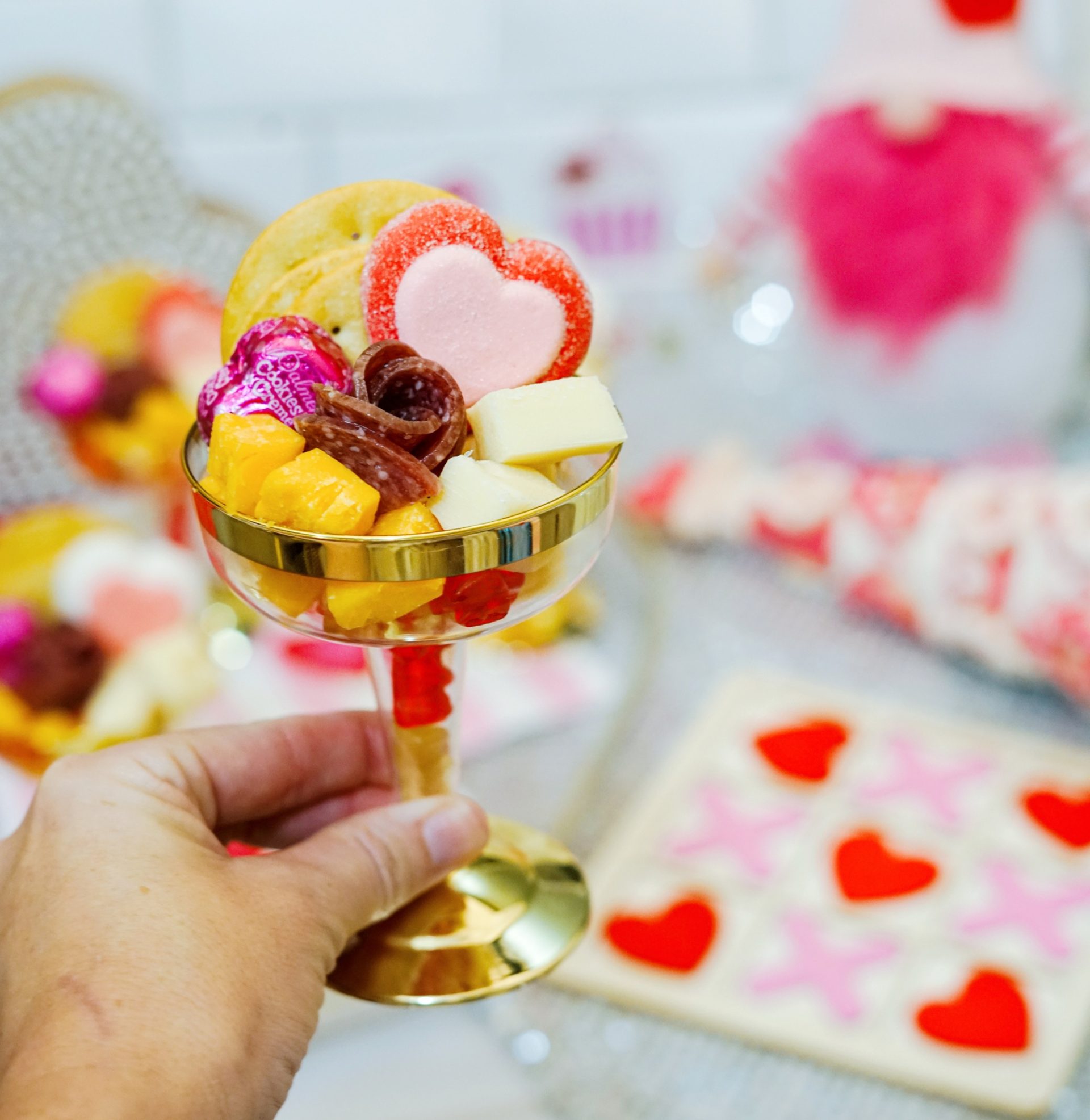 A person's hand holding a glass filled with Valentines charcuterie foods like cheese, candy, pepperoni flower and crackers.