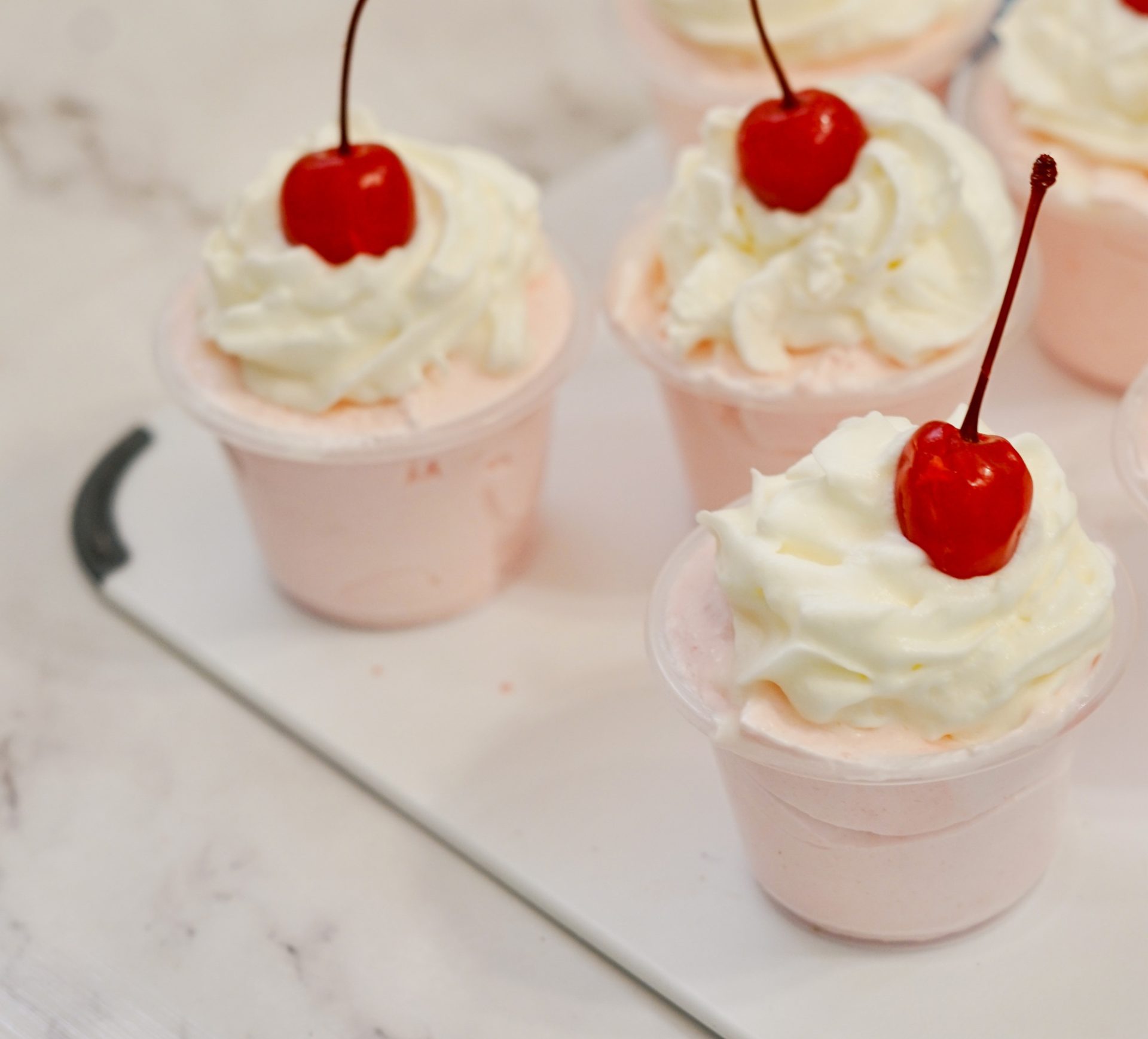 Pink tequila pudding shots with whipped cream and cherry garnish sit on a Galentines party table.