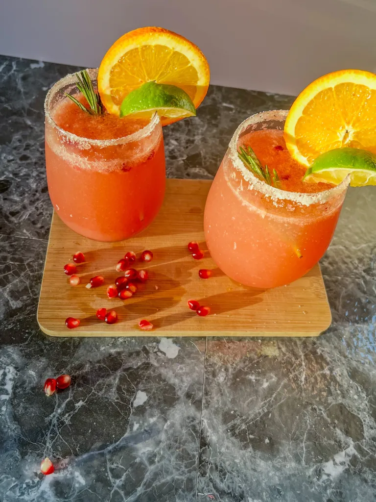 Orange and pomegranate mimosas in sugar rimmed glasses on a wood board.
