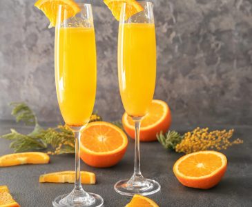 Mimosa cocktails sit on a table with oranges